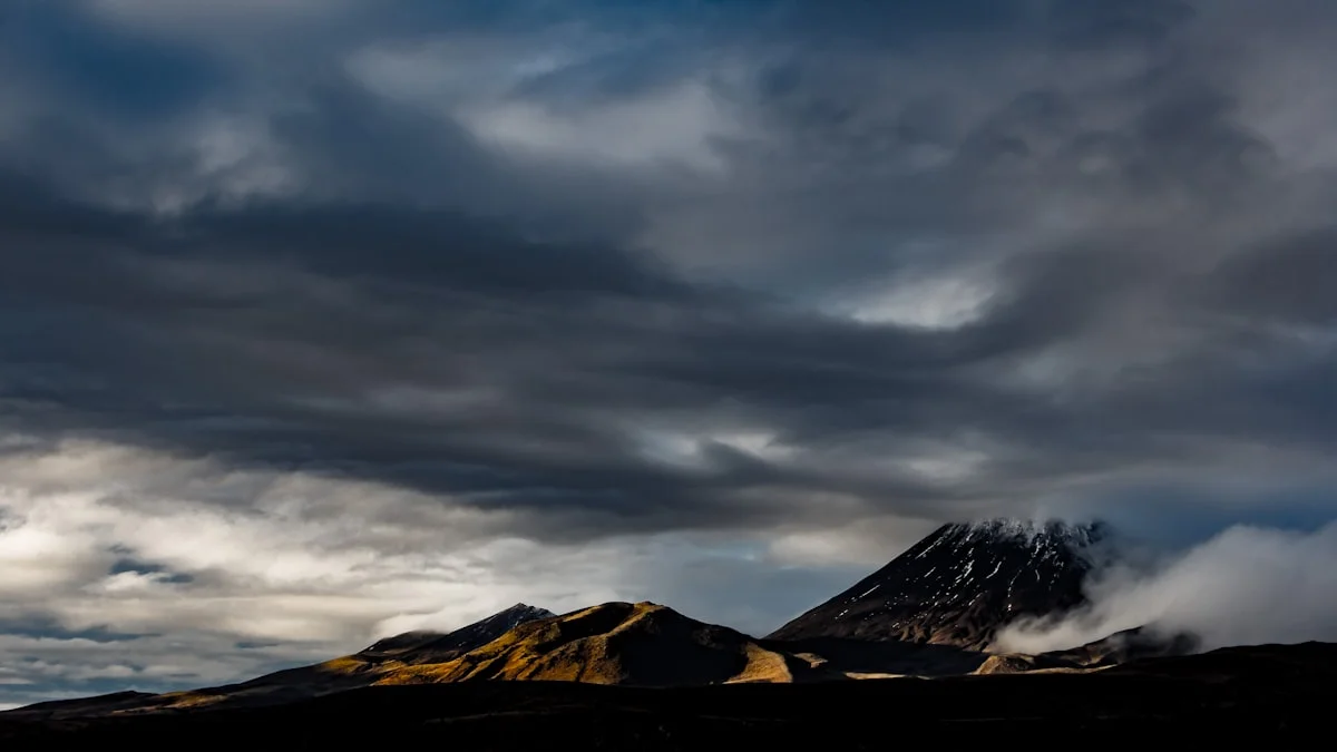Heavy grey clouds gather over dark mountain peaks with warm light on the ridges. — a The Obsidian Key guide to Precipitation: Why the Medium Makes You Wait.