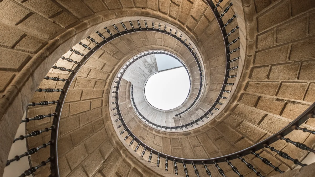 A worn stone spiral staircase winds upward toward a soft pale skylight. — a The Obsidian Key guide to Start Tonight: the Full Evening Protocol in Order.
